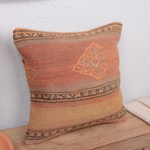 May include: A square decorative pillow with a woven design in shades of orange, peach, and tan. The pillow features a geometric pattern in the center and a patterned border. The pillow is placed on a wooden surface.