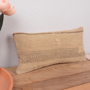 May include: A rectangular beige woven pillow with a darker brown stripe pattern. The pillow is placed on a wooden surface. Decorative flowers in a glass vase are in the background.
