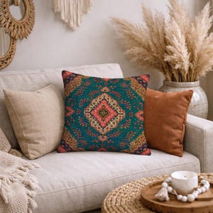 May include: A square throw pillow featuring a deep teal background with a central diamond-shaped medallion in shades of pink, cream, and gold, surrounded by intricate floral and vine patterns in a woven textile finish with dark blue edges.