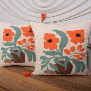 May include: Two white throw pillows with orange embroidered flowers and green leaves in a vase. The pillows have orange tassels.