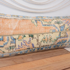 Pode incluir: Almofada decorativa retangular com um design de tapete vintage. A almofada apresenta um design de patchwork com uma parte superior laranja clara e uma parte inferior estampada com detalhes azuis, verdes e laranja. A almofada é colocada numa superfície de madeira.