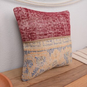 May include: A decorative pillow with a two-tone design. The top half is a deep red with a small white grid pattern, while the bottom half features a beige and blue patterned design. The pillow is placed on a wooden surface.