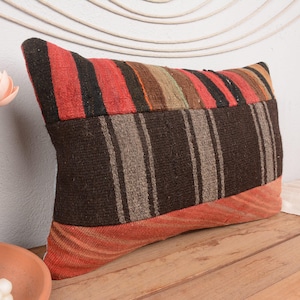 May include: Rectangular pillow with a patchwork design. The top features red and black stripes, transitioning to vertical brown and gray stripes. The bottom is a solid orange-red. The pillow is placed on a wooden surface.