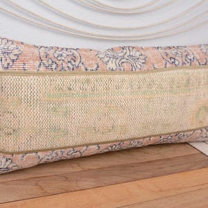 May include: Rectangular decorative pillow with a beige center and patterned borders in shades of pink and blue. The pillow is placed on a wooden surface, showcasing its textured fabric and design. A stylish home decor item.