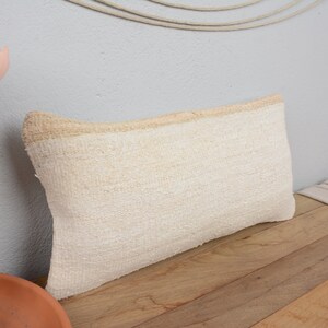 May include: A white throw pillow with a subtle texture and a cream-coloured border. The pillow is sitting on a wooden surface.