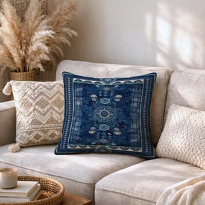 Puede incluir: Un cojín cuadrado con una base azul profundo y un patrón central de medallón intrincado en tonos azul claro, blanco y verde, enmarcado por un borde decorativo con motivos geométricos y una fina línea de acento en verde claro.