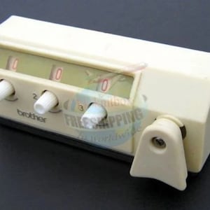 以下が含まれることがあります： 3つのダイヤルとレバーが付いた白いブラザーミシンフットコントローラー。ダイヤルには0、1、2、3の数字が書かれています。レバーには「brother」と書いてあります。