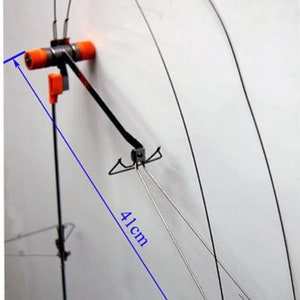Puede incluir: Un soporte para cañas de pescar de metal negro con un mango naranja y una altura de 41 cm. El soporte tiene un marco de alambre que se extiende hacia un lado.