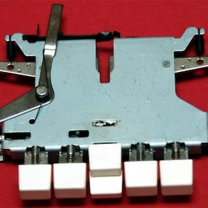 Op de afbeelding: Een vintage schrijfmachine-toetsmechanisme met een zilverkleurig metalen frame en witte vierkante toetsen. Het mechanisme omvat hendels, veren en andere kleine onderdelen. De toetsen zijn in een rij gerangschikt.