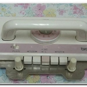 May include: White Brother KH-260 knitting machine with a pink control panel and a handle. The machine has a dial with numbers 1-10 and a row of buttons labeled "Plain", "Tuck", "=MO", "=L", "=R", and "Start".
