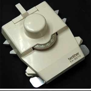 Könnte beinhalten: Cremefarbenes Brother KA-8210 Strickmaschinen-Zubehör. Es verfügt über ein zentrales Zifferblatt mit Zahlenmarkierungen, ein kleineres Zifferblatt und mehrere Metalllaschen. Das Gerät ist für die Verwendung mit einer Strickmaschine konzipiert.