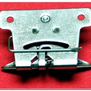 May include: Silver metal latch with a spring mechanism. The latch has two holes on the top and a small screw in the center.