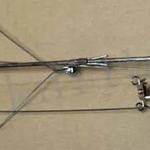 Puede incluir: Un devanador de hilo de metal plateado con una bobina de plástico blanco. El devanador tiene un brazo largo con un gancho en el extremo y un brazo más pequeño con una perilla de tensión. El devanador se utiliza para enrollar el hilo en una bola.