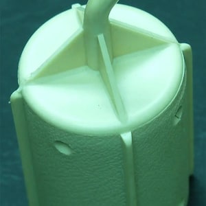 May include: A pale yellow plastic hook with a cylindrical base. The hook is curved and extends from the top of the cylinder. The base has a cross-shaped design on top and vertical supports along the sides. There are oval-shaped holes on the sides.