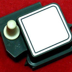 May include: A dark gray plastic component with a white square and a beige knob. The square has a black border. The component is on a red surface. The knob is cylindrical with a ridged texture.