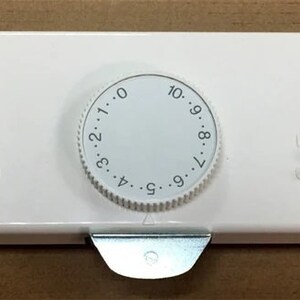 May include: A white thermostat with a circular dial numbered 0-10. The device has a rectangular shape with two sets of clips on either side and a metal tab at the bottom. The thermostat is designed for temperature control.