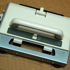 May include: Cream-colored knitting machine with a silver metal base and a white handle. The machine has a dark green panel with the letters "N" and "F" and the letter "L". The machine is designed for knitting.