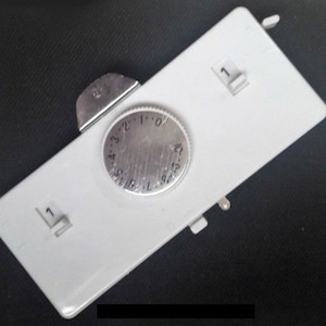 May include: A rectangular, light gray control panel with a central, circular dial and two small switches. The dial is silver with numbers from 0 to 10. The switches are labeled with the number 1. The panel has a metallic lever.