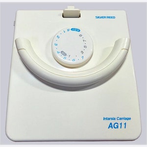 May include: White Intarsia Carriage AG11 by Silver Reed with a dial for adjusting the tension. The dial has numbers 1 through 9 and a "6.5" marking.
