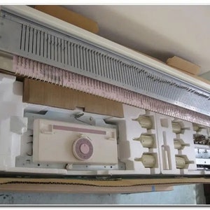 May include: A white knitting machine with a gray needle bed and a pink yarn holder. The machine is in a cardboard box with styrofoam packing.