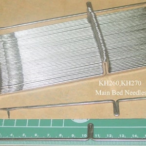 May include: A collection of silver-colored main bed needles for a knitting machine. The needles are arranged in a stack and individually. The text "KH260, KH270 Main Bed Needles" is visible. A ruler is in the foreground.