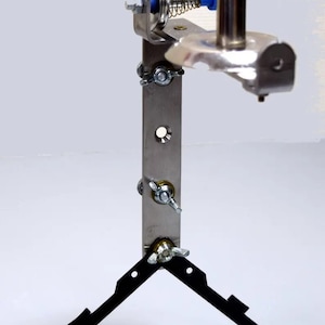 Puede incluir: Un soporte para cañas de pescar de metal plateado con una base de trípode negra. El soporte tiene un brazo con resorte con una rueda giratoria y una perilla para ajustar la tensión.