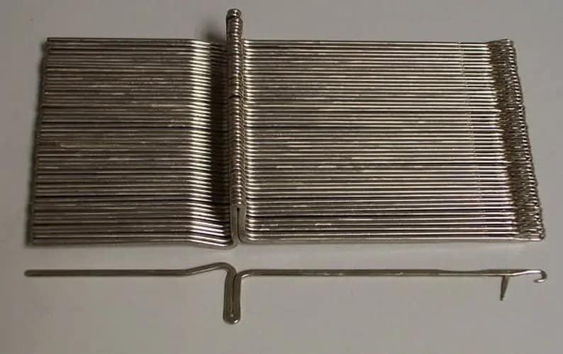 K&ouml;nnte beinhalten: Ein Satz silberner Stricknadeln mit einer einzelnen Nadel f&uuml;r einen Latch-Hook. Die Nadeln sind in einem Stapel angeordnet und die Latch-Hook-Nadel liegt unter dem Stapel.