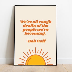 May include: A beige poster with a black frame features a quote in orange text: "We're all rough drafts of the people we're becoming." - Bob Goff. The poster has a stylised orange sun with yellow rays at the bottom.