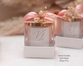 May include: Two small glass candle jars with gold lids and white labels. The labels have the initials "VL" and the text "Victoria Lina". The jars are decorated with pink ribbon and dried flowers. The jars are sitting on white boxes with the text "Somon Kurdele, Gold Kapak, Beyaz Kutu".