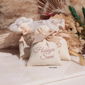 May include: Two small white fabric bags with lace trim and gold ribbon tied around them. The bags are decorated with the names "Husseyn" and "Sami" in pink script. The text "Gold Kurdele" is printed on the bottom of the bag.