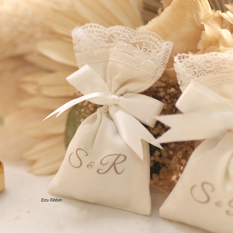 May include: Two small white fabric bags with lace trim and a white satin ribbon tied in a bow. The bags are embroidered with the initials "S&R" and "S&S" in brown thread. The bags are sitting on a background of dried flowers.