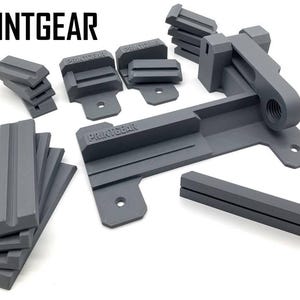 Könnte beinhalten: Ein Satz grauer 3D-gedruckter Teile für einen Drucker, bestehend aus einer Basis, einem Hebel und mehreren kleineren Teilen. Die Teile sind mit "PRINTGEAR" beschriftet.