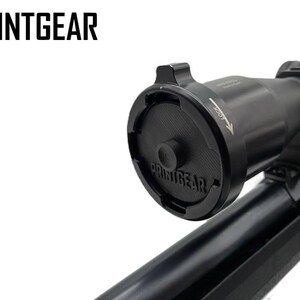Könnte beinhalten: Schwarzes Zielfernrohr mit PRINTGEAR-Logo und Schutzabdeckung. Das Zielfernrohr ist auf einem schwarzen Gewehr montiert, vor weißem Hintergrund. Das Zielfernrohr hat Einstellräder und Markierungen.