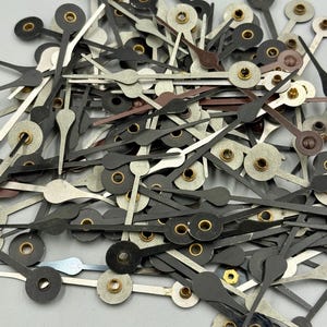 May include: A pile of clock hands in various colors, including black, silver, and brown. The hands have a variety of shapes and sizes, with gold-colored metal inserts. The hands are scattered, suggesting a collection of watch or clock repair parts.