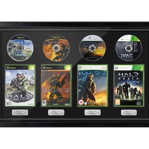 May include: A framed collection of four Halo video game discs and their covers. The black frame displays the game discs and their cases. The game cases feature soldiers and futuristic scenes. The discs are labelled with the Halo logo.