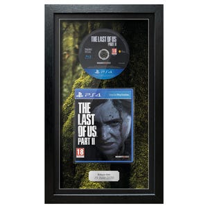 May include: Framed artwork featuring a PlayStation 4 game disc and game case for "The Last of Us Part II." The disc is above the game case, both set against a mossy background. The game case features a portrait of a woman.