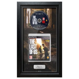May include: Framed PlayStation 3 game and disc for "The Last of Us." The game case and disc are displayed in a black frame. The game case features a sunset scene with two figures. The disc has the game title and an age rating.