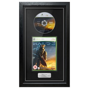 Op de afbeelding: Ingelijste Halo 3 Xbox 360 game en disc. De game case toont een futuristische soldaat. De disc bevindt zich boven de game case. De lijst is zwart met een zilveren plaatje onderaan: "Release Date 25 September 2007."