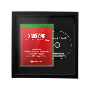 May include: A black framed display featuring an Xbox One game case and a disc. The game case has a red and green design with the text "XBOX ONE" and "USE YOUR OWN GAME". The disc is labeled "FRAME-A-GAME".