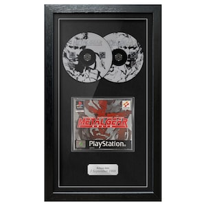 May include: Framed Metal Gear Solid PlayStation game and two discs. The game case features red and gray graphics with the title. The discs have a black and white design. The frame is black with a white matting.