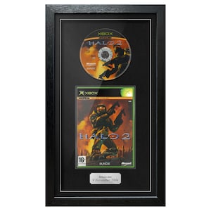 May include: Framed Halo 2 Xbox game and disc. The game case features a soldier with weapons against a fiery background. The disc and case are displayed in a black frame with a release date plaque.