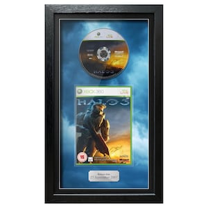 May include: Framed Halo 3 Xbox 360 game and disc. The game case features a warrior figure against a sunset backdrop. The disc is above the game case. The frame is black with a blue background.