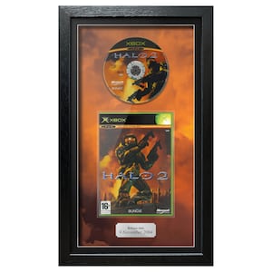 May include: Framed Halo 2 Xbox game and disc. The game case features a soldier with weapons, and the disc shows a similar design. The frame includes a release date of November 9, 2004.