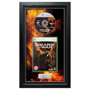 May include: Framed Gears of War Xbox 360 game and disc. The game case features a soldier with a weapon, set against a fiery background. The disc has the game title and a skull graphic. The frame is black.