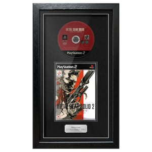 May include: Framed Metal Gear Solid 2 video game display. The black frame holds a red PlayStation 2 disc and the game case. The game case features a man with a gun. The release date is November 13, 2001.