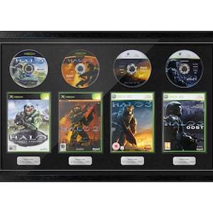 May include: Framed collection of four Halo video game discs and game covers. The black frame holds the discs and game cases, showcasing the game titles and release dates. The games are for Xbox and Xbox 360.
