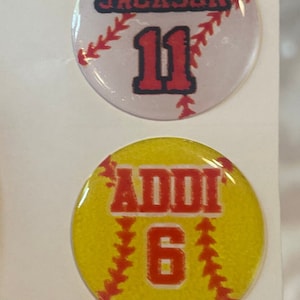 May include: Four round stickers with a softball design. The top two stickers are white with red stitching and the text "JACKSON 11". The bottom two stickers are yellow with red stitching and the text "ADDI 6".