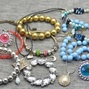 Op de afbeelding: Een verzameling kettingen en armbanden met verschillende kralen, bedels en hangers. De sieraden omvatten een zilveren ketting met een rozenhanger, een roze kralenarmband, een gouden kralenarmband, een blauwe kralenketting en een zilveren ketting met een kristallen hanger.