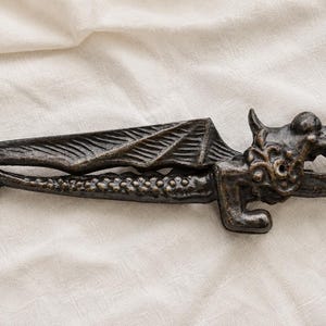 May include: A dark bronze-coloured metal nutcracker shaped like a dragon with a textured wing and scaled body. The dragon's head features an open mouth with visible teeth. A clear plastic ruler is positioned to the left, showing a length of 30 cm.