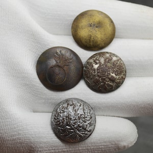 Op de afbeelding: Vier antieke metalen knopen met verschillende ontwerpen. Eén knoop is goud, één is zilver en twee zijn brons. De knopen hebben verschillende ontwerpen, waaronder een granaat, een wapenschild en een bloemenpatroon.